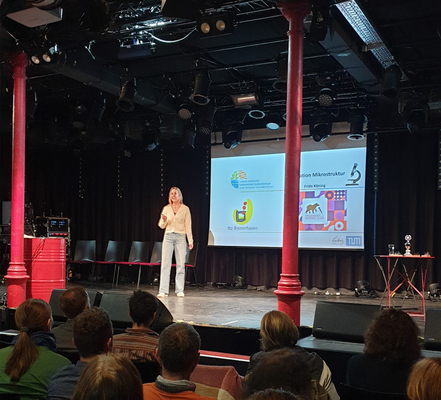 Frida König hält ihren Vortrag auf der Slammer-Bühne. Sie trägt eine hellblaue Jeans und ein sandfarbenes Oberteil. Im Hintergrund ist eine Präsentationsfolie ihres Vortrags zu sehen.