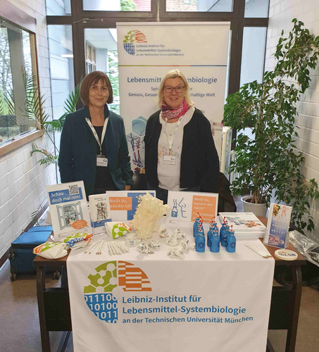 Hier sehen Sie den Infostand des Leibniz-Instituts für Lebensmittel-Systembiologie an der Technischen Universität München in einem Innengang der Chemieschule. Hinter einem Tisch mit Broschüren, Informationsmaterialien, einem 3D-Rezeptormodell, Geruchsproben in blauen Fläschchen und Werbeartikeln stehen zwei Frauen. Hinter dem Stand befindet sich ein Roll-up-Banner mit dem Logo des Instituts und Informationen zur Lebensmittel-Systembiologie. Seitlich rahmen Pflanzen den Stand ein.