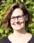 Portraitfoto von Kim J. Herrmann, sie hat mittellanges dunkelbraunes Haar und trägt eine Brille, sie steht vor einem grünbelaubten Busch.