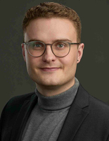 Portrait photo of Phil Richter, who has short brown hair, wears glasses, and dark clothing. The background is dark.