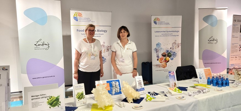 Präsentationsstand des Leibniz-LSB@TUM mit Exponaten, Informationsmaterialien und Roll-ups zum Forschungsthema nachhaltige Proteinquellen auf den Münchner Wissenschaftstagen 2026. Hinter den Infotischen des Stands stehen zwei Standbetreuerinnen und schauen freundlich in die Kamera. Sie tragen weiße Oberteile.