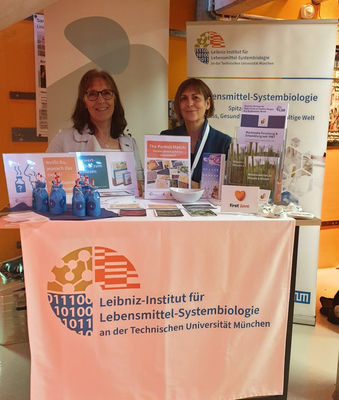 Inge Kirchmann (links) und Dr. Gisela Olias stehen hinter einem Tisch, auf dem verschiedene Exponate zu sehen sind. Im Vordergrund ist auf der Tischdecke das Logo des Leibniz-Instituts zu sehen. Im Hintergrund sind Roll-ups zu sehen. Auf dem rechten Roll-up ist das Logo des Leibniz-LSB@TUM zu sehen.